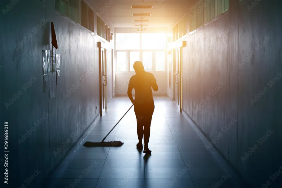 Stock photo of a custodian pushing a wide broom down a sunlit hallway
